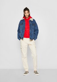 Denim jacket with a shearling collar, red hoodie underneath, beige cargo pants, and black-and-white checkered slip-on shoes.