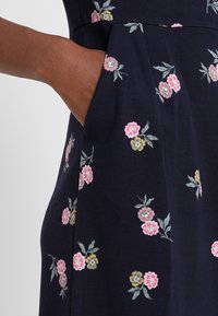 Marinblå klänning med broderade blommönster i rosa, gult och grönt. Har sidofickor och en figurnära midja med veck.