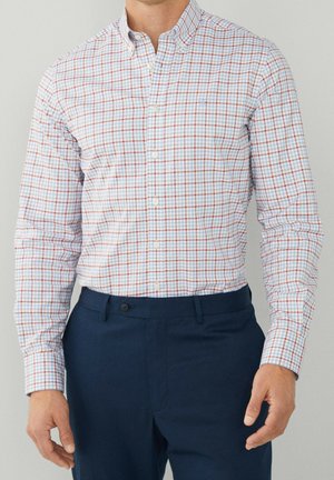 Homme portant une chemise à carreaux bleu clair, rouge et blanc à manches longues, rentrée dans un pantalon bleu foncé, debout les bras détendus.