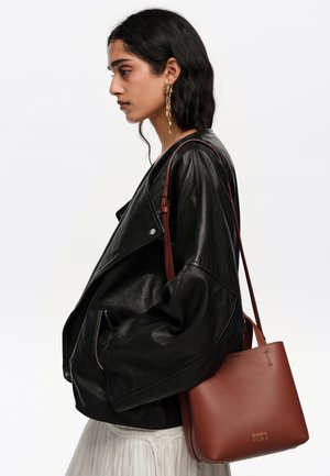 Mujer con chaqueta de cuero negra llevando un bolso de cuero marrón sobre el hombro, con pendientes largos dorados y falda blanca.