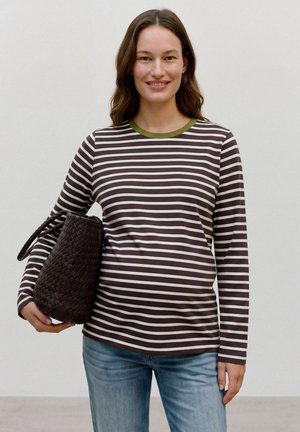 Femme souriante en chemise à rayures manches longues et jean tenant un grand sac à main tissé marron foncé devant un fond uni.