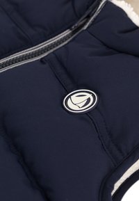 Temno modra puhovka brez rokavov s sijočo teksturo, z zadrgo spredaj in OKROGLIM logotipom. Mehka, bela podloga na ovratniku.