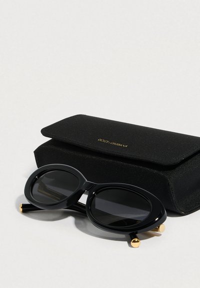 Μαύρα γυαλιά ηλίου με οβάλ φακούς και χρυσές λεπτομέρειες, συνοδευόμενα από μαύρη θήκη με υφή και μαγνητικό κλείσιμο.