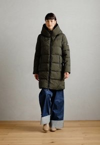 Olijfgroene pufferjas met een hoge capuchon, ritsluiting en horizontale stiksels. Gecombineerd met denim jeans met wijde pijpen en beige schoenen.