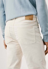 Pantaloni di velluto bianco con tasche posteriori e un'etichetta in pelle marrone. Il modello indossa un maglione azzurro chiaro con polsini a coste.