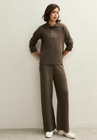 Pull en knit marron avec un col et trois boutons, associé à un pantalon large marron. Texture lisse, coupe décontractée, complété par des baskets blanches.