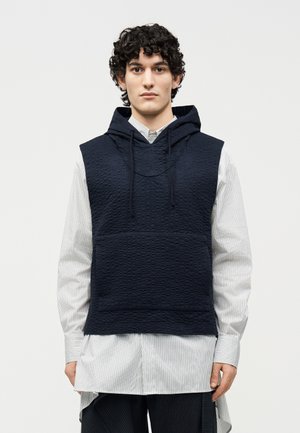 Νεαρός άνδρας με σγουρά μαλλιά, φοράει μια υφασμάτινη μπλε σκούρα αμάνικη φούτερ με κουκούλα πάνω από ένα μακρύ, ανοιχτόχρωμο ριγέ πουκάμισο και σκούρο παντελόνι, σε λευκό φόντο.