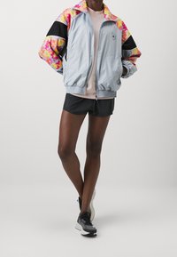 Chaqueta cortavientos de color gris claro con acentos vibrantes en rosa, naranja y amarillo, mangas negras, puños elásticos y cierre con cremallera en la parte delantera.