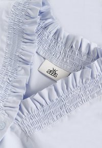 Hele sinine pluus, millel on volangiga krae ja smocking detailid. Sisaldab brändi sildit, millel on stiliseeritud logo. Sile kangastekstuuri.