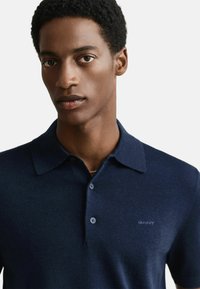 Mladý muž s krátkými kudrnatými vlasy, oblečený v námořnicky modrém polokošili s límečkem, se dvěma knoflíky a diskrétním logem na prsou.