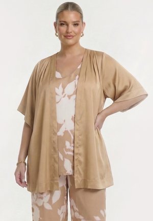 Vrouw met gouden hoepeloorbellen die een losse tan-kleurige korte mouwen open jas draagt over een beige-wit gebloemde top en bijpassende broek, hand op de heup.