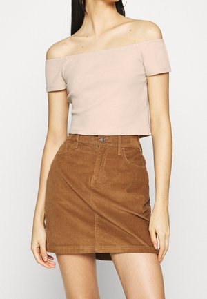 Vrouw die een beige off-shoulder geribbelde crop top en een high-waisted bruine corduroy minirok draagt, staand tegen een effen achtergrond.