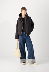 Veste noire matelassée avec capuche, fermeture éclair, portée avec un jean en denim bleu à jambes larges et un sac beige. Design simple sans motifs.