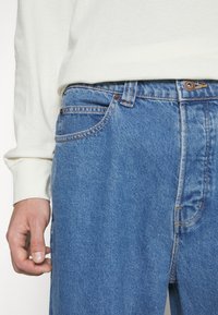 Light blue denim jeans with a relaxed fit. Features contrast stitching, a button closure, and front pockets. Worn with a white long-sleeve shirt.