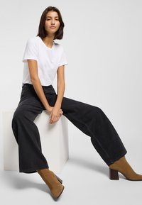 Young woman sits on white cube wearing white t-shirt, wide black jeans, and brown heeled ankle boots against a plain light gray background.