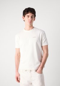 Baltas trumpas rankovėmis marškinėliai, pagaminti iš lygaus audinio. Yra diskretus Calvin Klein logotipas ant krūtinės. Paprastas, švarus dizainas be raštų.