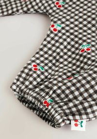 Barna és fehér kockás textil, amelynél ráncolt mandzsetta található, piros cseresznye hímzéssel és zöld levelekkel, kicsi cseresznye címkével díszítve.