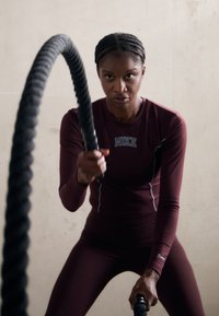 Maroon välsittande träningslinne och leggings med Nike-logotyp. Individ som greppar ett tjockt träningsrep, med ett fokuserat utseende, enkel bakgrund.