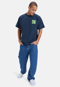 Marineblå bomull T-skjorte med en dristig grønn grafisk logo på brystet, kombinert med lyseblå løstsittende denimjeans og hvite joggesko.
