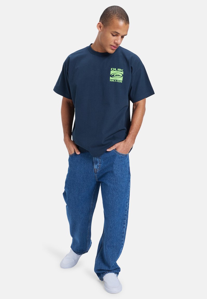 Marineblå bomull T-skjorte med en dristig grønn grafisk logo på brystet, kombinert med lyseblå løstsittende denimjeans og hvite joggesko.