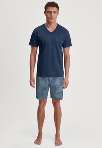 Jonge man die blootsvoets staat, gekleed in een marineblauwe V-hals shirt en blauwe, met een patroon bedekte knielange shorts, tegen een witte achtergrond.
