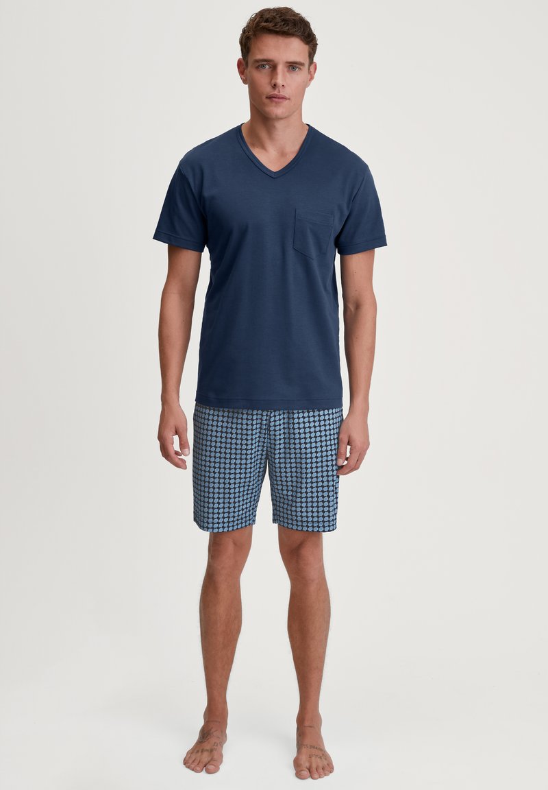 Jonge man die blootsvoets staat, gekleed in een marineblauwe V-hals shirt en blauwe, met een patroon bedekte knielange shorts, tegen een witte achtergrond.