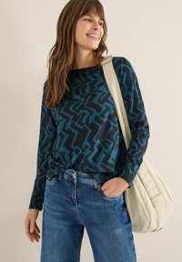 Langarmtop in donkerblauw met abstracte turquoise patronen, gecombineerd met lichtblauwe jeans. Het model draagt een schoudertas in crèmekleur.