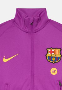 Violeta sporta jaka ar augstu apkakli, kurai ir dzeltenais Nike swoosh, izšūts FC Barcelona logo un Spotify ikona.
