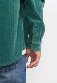 Donkergroen corduroy shirt met ribbeltextuur, knoopmanchetten en een afgeronde zoom, gecombineerd met blauwe denim jeans.