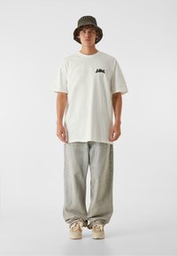 Tricou alb din bumbac cu mâneci scurte, grafică mică neagră pe piept, combinat cu pantaloni oversized din denim gri deschis și adidași bej.