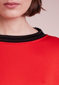 Sudadera roja con un cuello de canalé negro en contraste, que presenta sutiles acentos de rayas doradas. Textura suave con una vista en primer plano del escote.