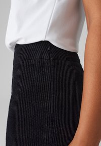 Black corduroy skirt with vertical ridges, featuring a concealed zipper at the back. Paired with a white shirt. Side view.
