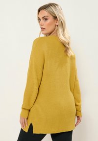 Pull en maille moutarde à manches longues, texture côtelée, fentes latérales et coupe décontractée, assorti à un pantalon noir.