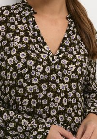 Blouse florale en tissu vert foncé avec des fleurs blanches et violettes. Elle présente un col en V et des manches longues avec des fronces aux poignets.