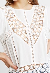 Blusa bianca con motivi di pizzo circolari, inserti trasparenti e una vestibilità morbida. Presenta cuciture dettagliate e un tessuto strutturato.