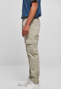 Beige cargo bukser med sidelommer, elastisk talje og smalle ankler, kombineret med hvide sneakers og en blå t-shirt, vist fra siden.