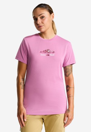 Frau trägt ein pinkes T-Shirt mit Berggrafik und "Everest Massif" Text, beige Hose und sichtbare Tattoos auf beiden Armen.
