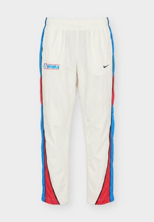 Homme portant un pantalon de basket WNBA blanc avec des bandes latérales rouges et bleues, logos Nike et baskets blanches, se tenant devant un fond neutre.