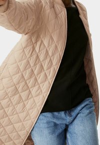 Vadderad, ljusrosa jacka med dragkedja och ribbad krage. Bärs över en svart topp, med blå jeans synliga nedtill.