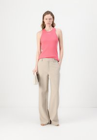 Rosa ermeløs topp med logo, sammen med lyse beige vid bukser. Modellen holder en liten kremfarget håndveske. Enkel, ren bakgrunn.