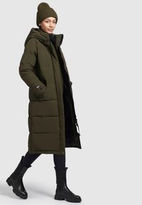 Lang olivengrøn puffjakke med hætte, der har lynlåslukning, sidelommer og sort foring. Sammen med sorte ankelstøvler.