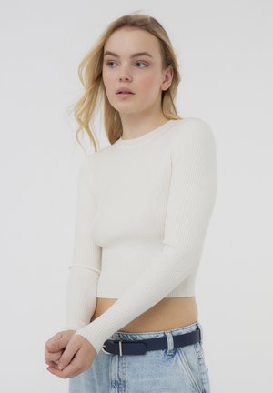 Top bianco a maniche lunghe con design aderente, scollo a girocollo e orlo a taglio, abbinato a pantaloni corti in denim blu a vita alta e una cintura nera.
