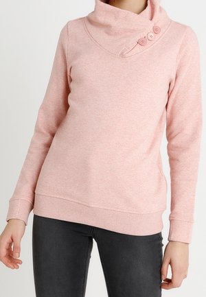 Vrouw draagt een lichtroze lange mouw pullover met een hoge kraag met twee knopen, gecombineerd met donkergrijze broek.