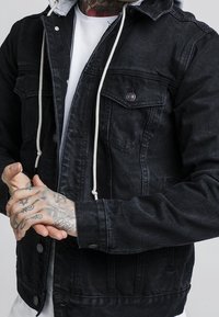 Schwarze Jeansjacke mit einer hellgrauen Kapuze, verstellbaren Zugbändern, Druckknopfverschluss und zwei Fronttaschen mit Klappen. Strukturiertes Gewebe.