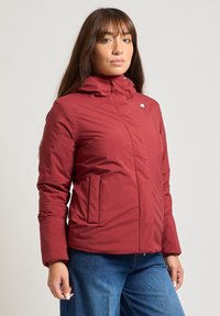 Giacca isolante rossa con chiusura a zip, colletto alto e due tasche frontali. Presenta un semplice badge con logo sul lato sinistro del petto.