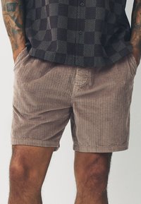 Cordhosen-Shorts in Hellbraun mit vertikaler Rippenstruktur. Verfügen über Seitentaschen und eine lockere Passform, kombiniert mit einem gemusterten, kurzärmligen Hemd.