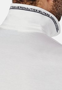 Polo shirt in cotone bianco con colletto alto. Il colletto presenta un logo ricamato blu con il testo. Texture liscia senza motivi visibili.
