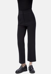 Mujer con pantalones negros de cintura alta y pierna recta con bolsillos delanteros, y loafers negros de tacón ancho, de pie contra un fondo liso.