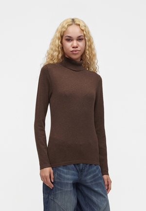 Jeune personne aux longs cheveux blonds bouclés portant un pull à col roulé marron et un jean bleu ample, debout devant un fond blanc.