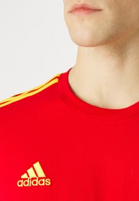 Rode t-shirt met een ronde hals. Heeft gele strepen op de schouders en een geborduurd geel Adidas-logo op de borst.